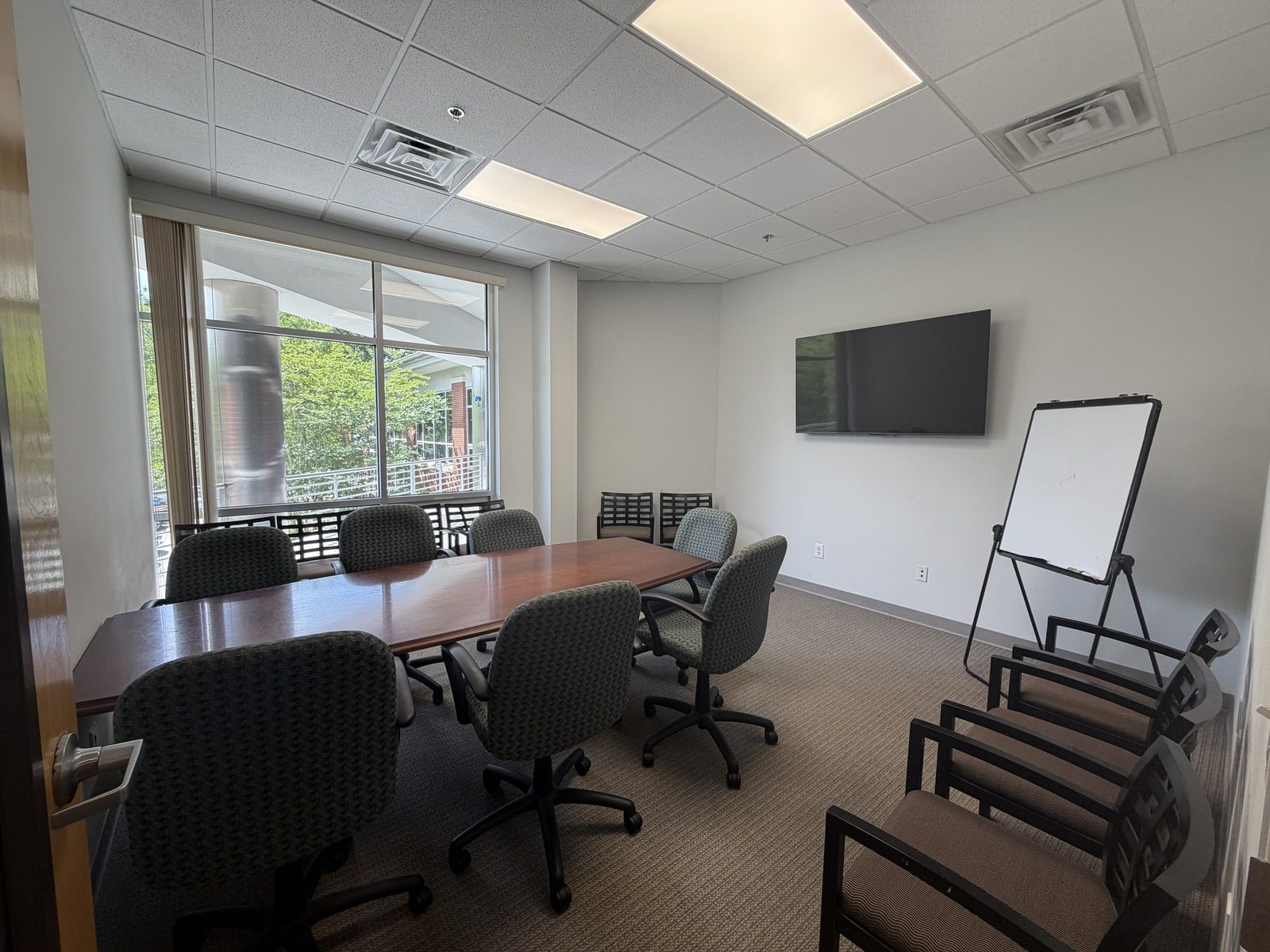 Conference room at Charleston West with a large wood table, twelve chairs, wall-mounted display, whiteboard, and window overlooking greenery